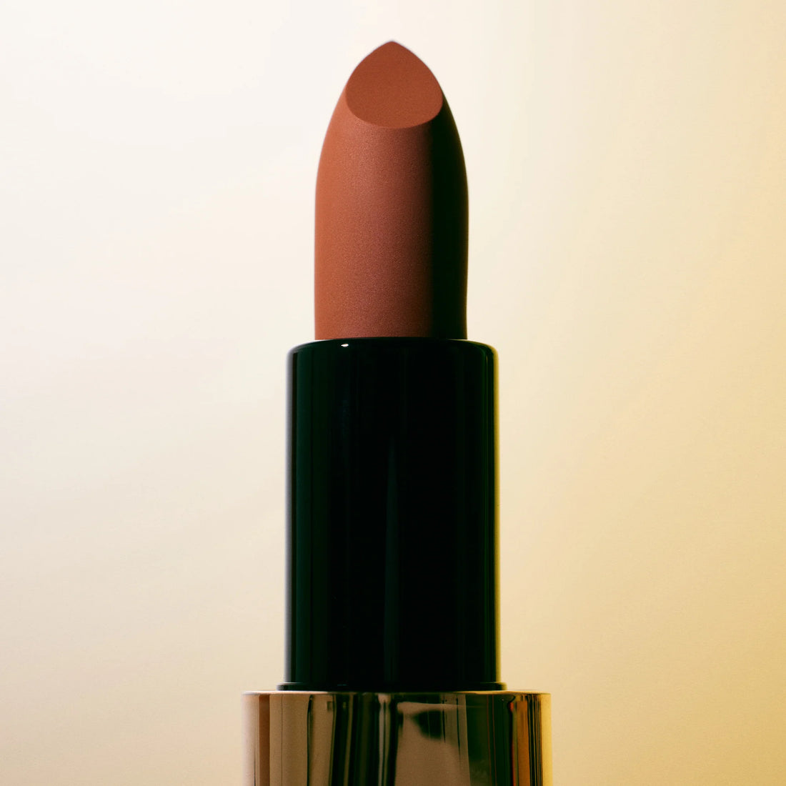 A lipstick shown open with the bullet exposed, alongside its cap placed beside it. The lipstick bullet is a warm medium brown, and the packaging features a metallic silver top, a black middle section, and a champagne-gold lower base.