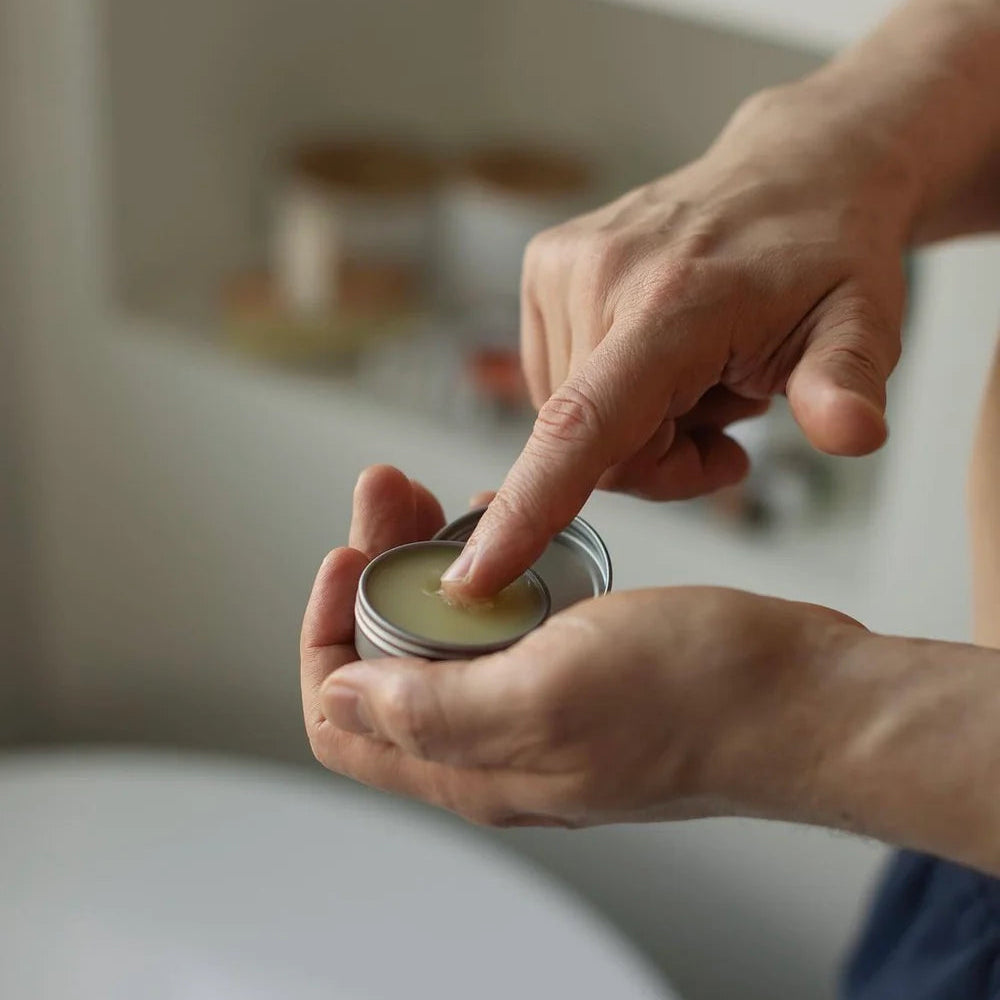 A close-up of a person's hands holding an open silver tin of balm or lip balm, with their finger touching the cream inside, against a softly blurred bathroom background.