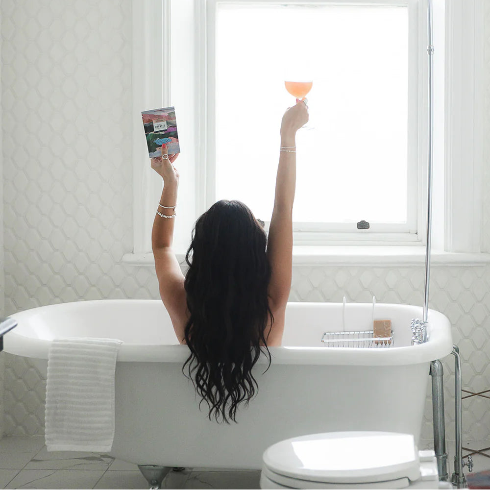 Person sitting in a bathtub with a book and a glass, looking out a window.
