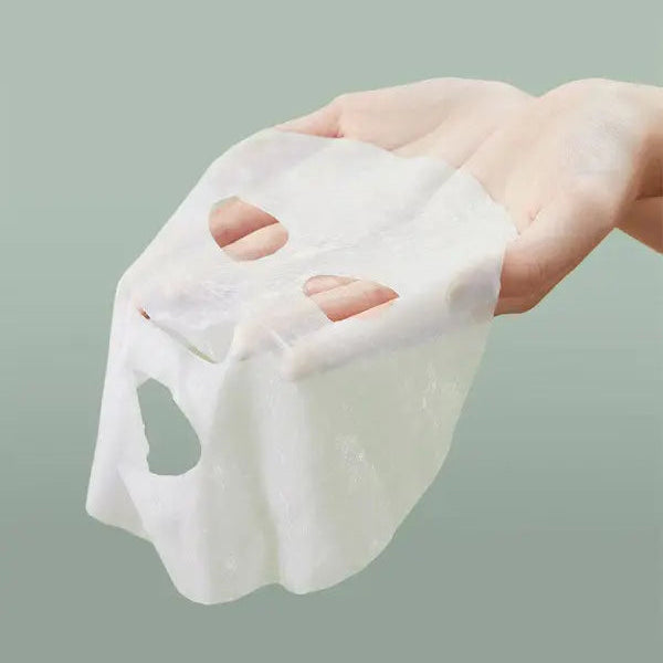 Hand holding a white sheet mask against a light green background