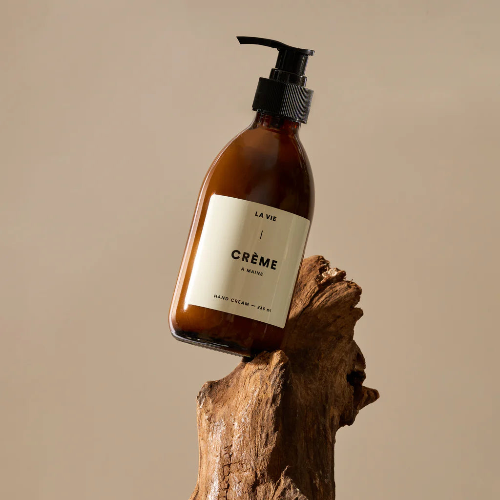 Hand cream bottle labeled 'La Vie Crème   Mains' on a wooden block with a beige background