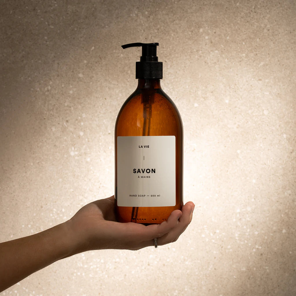 Hand holding an amber glass bottle labeled 'Savon' against a textured beige background