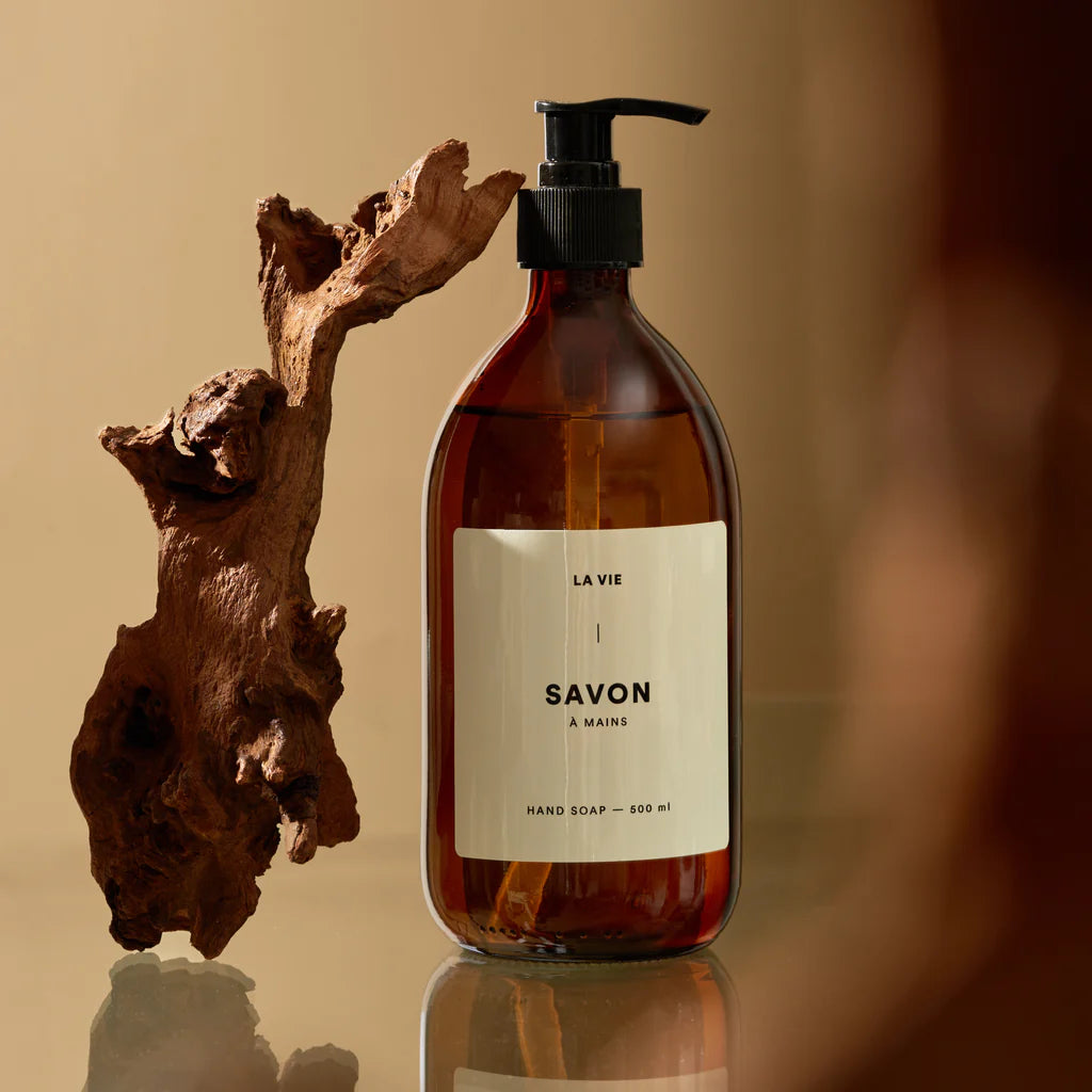 Brown soap bottle with pump and 'La Vie a Mains' label on a reflective surface with a wooden piece.
