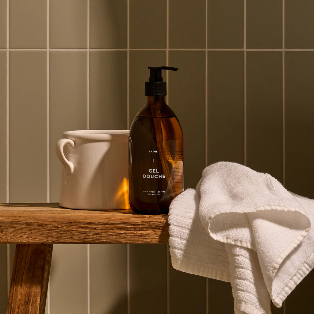Brown bottle labeled 'Gel Douche' on a wooden shelf with a white towel and ceramic cup against a tiled wall.