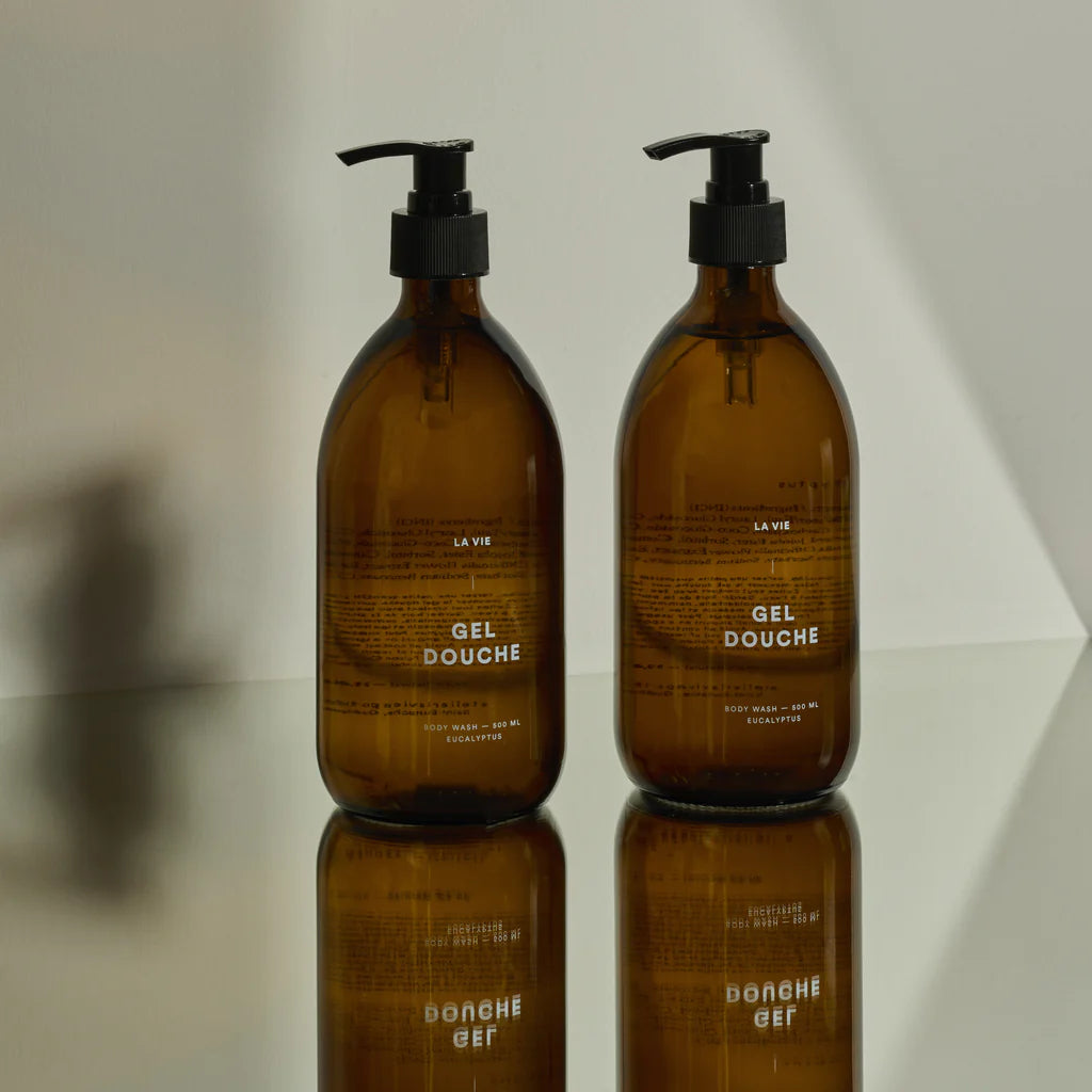 Two brown bottles labeled 'Gel Douche' on a reflective surface with a neutral background