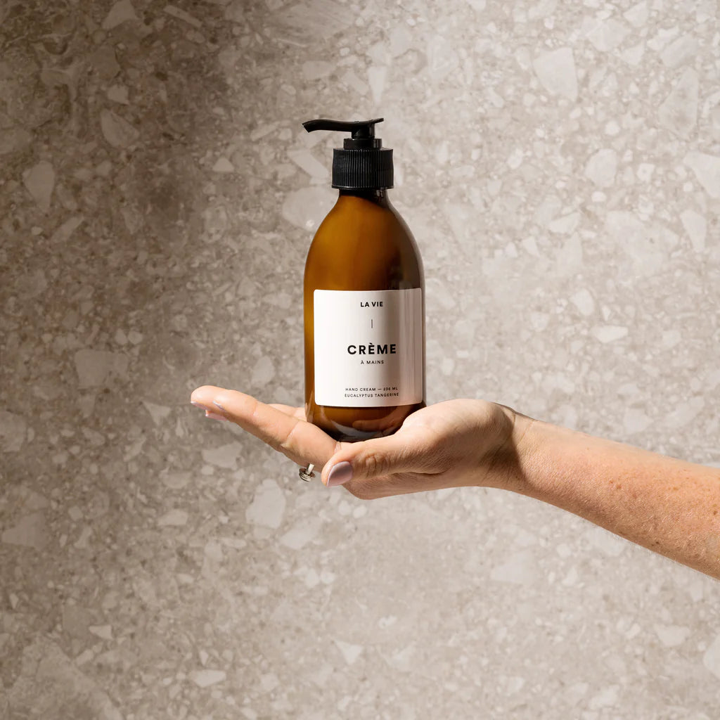 Hand holding a bottle labeled 'La Vie Crème' against a textured beige background