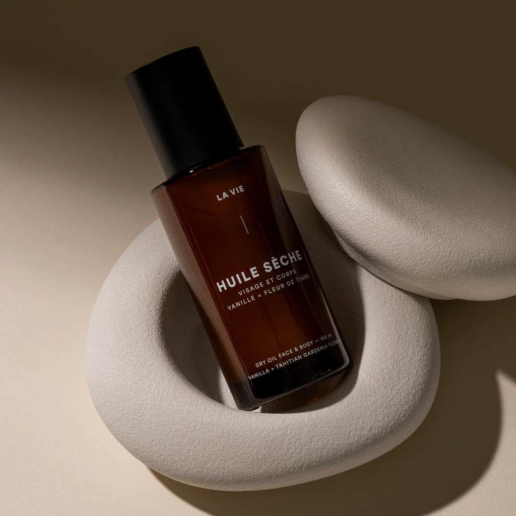 Brown bottle labeled 'Huile Seche' by LAVIE on a beige stone surface with a soft focus background.
