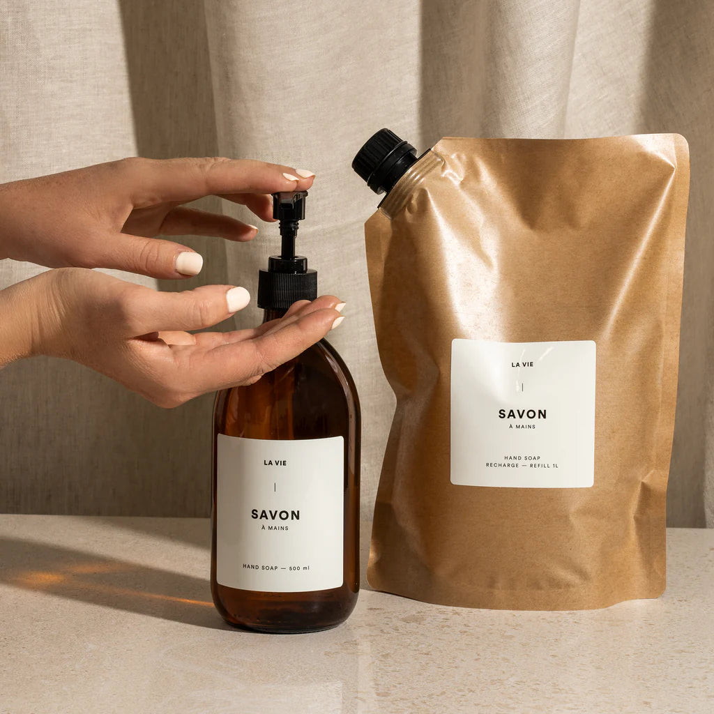 Hand holding a bottle of soap next to a brown paper bag with 'La Vie Savon' branding on a neutral background.