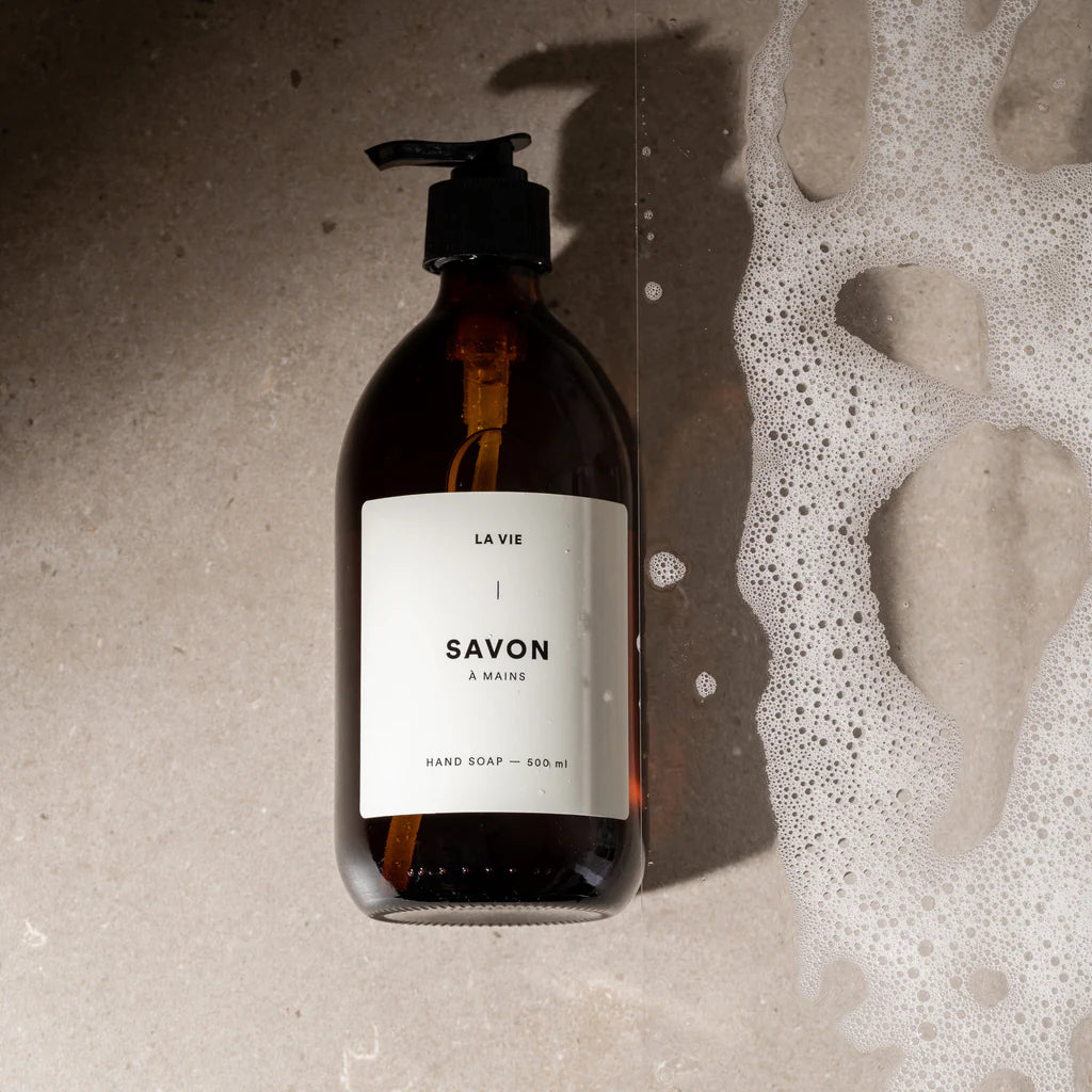 Hand soap bottle labeled 'SAVON' with a pump on a textured surface with soap suds.