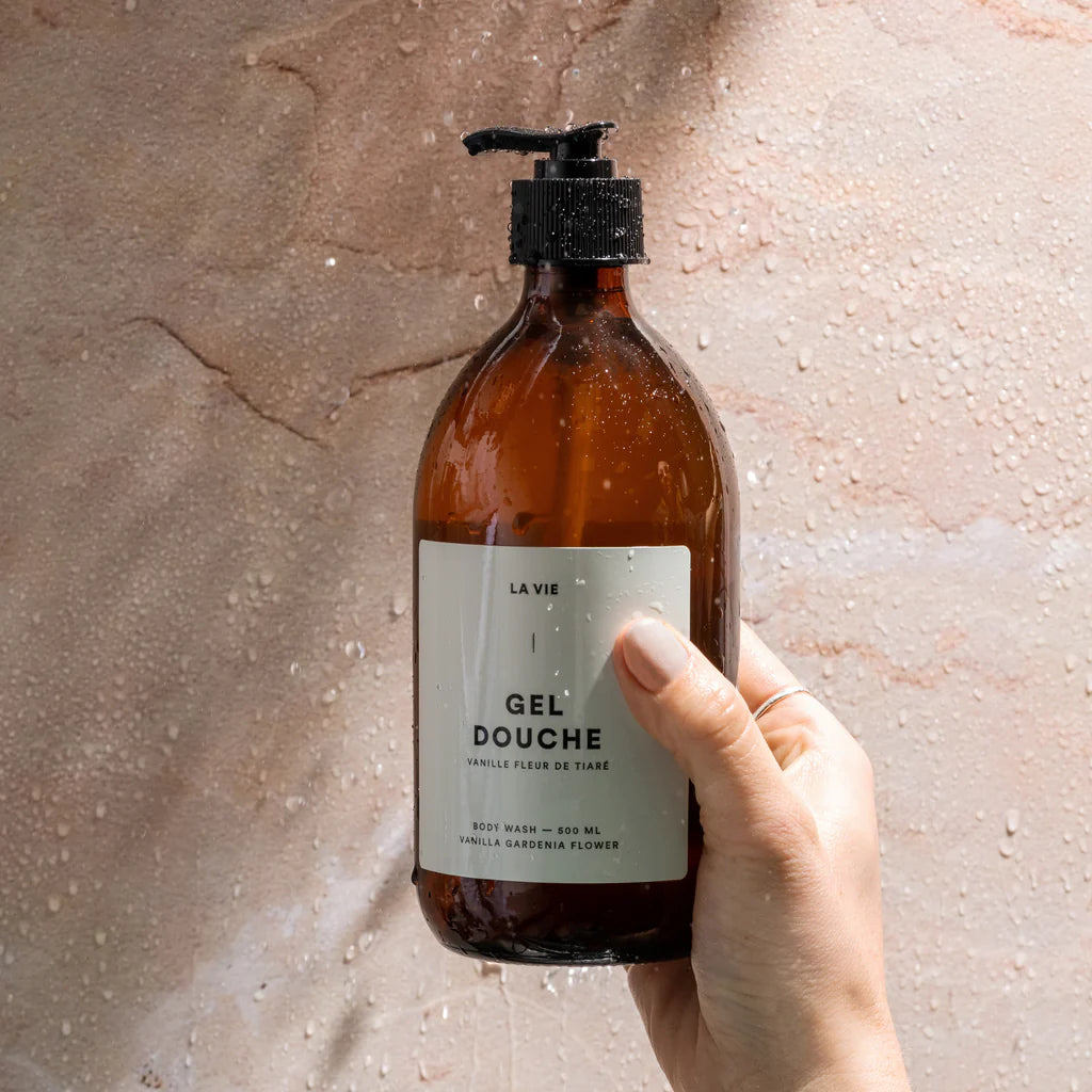 Hand holding a bottle of 'Gel Douche' with a label against a textured beige background