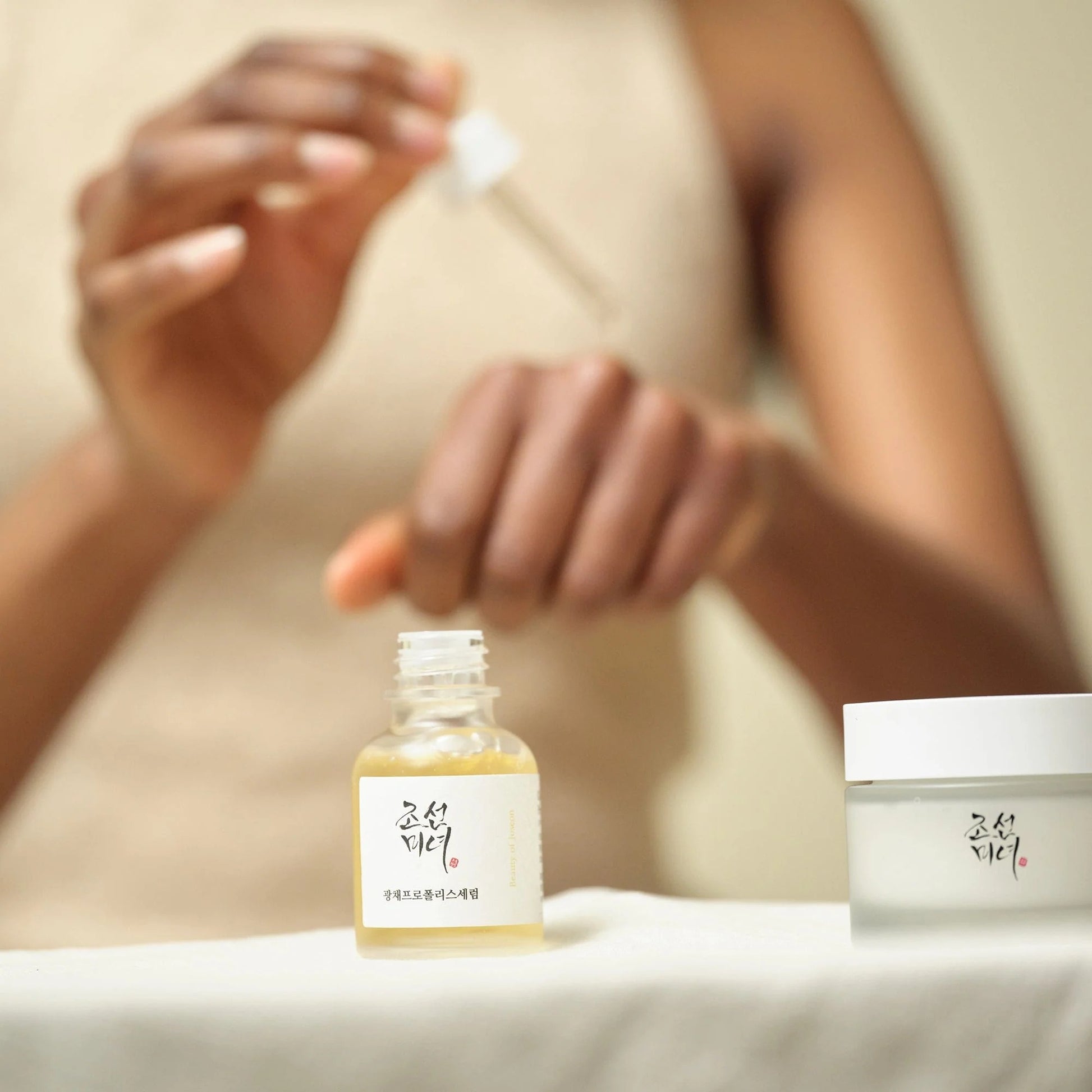 Person applying a skincare product with a dropper to a bottle and jar on a neutral background