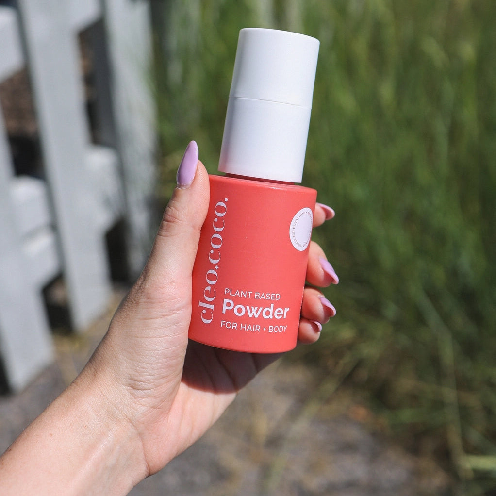 Hand holding a red and white bottle labeled 'cleo + coco plant based powder for hair + body' with a blurred background