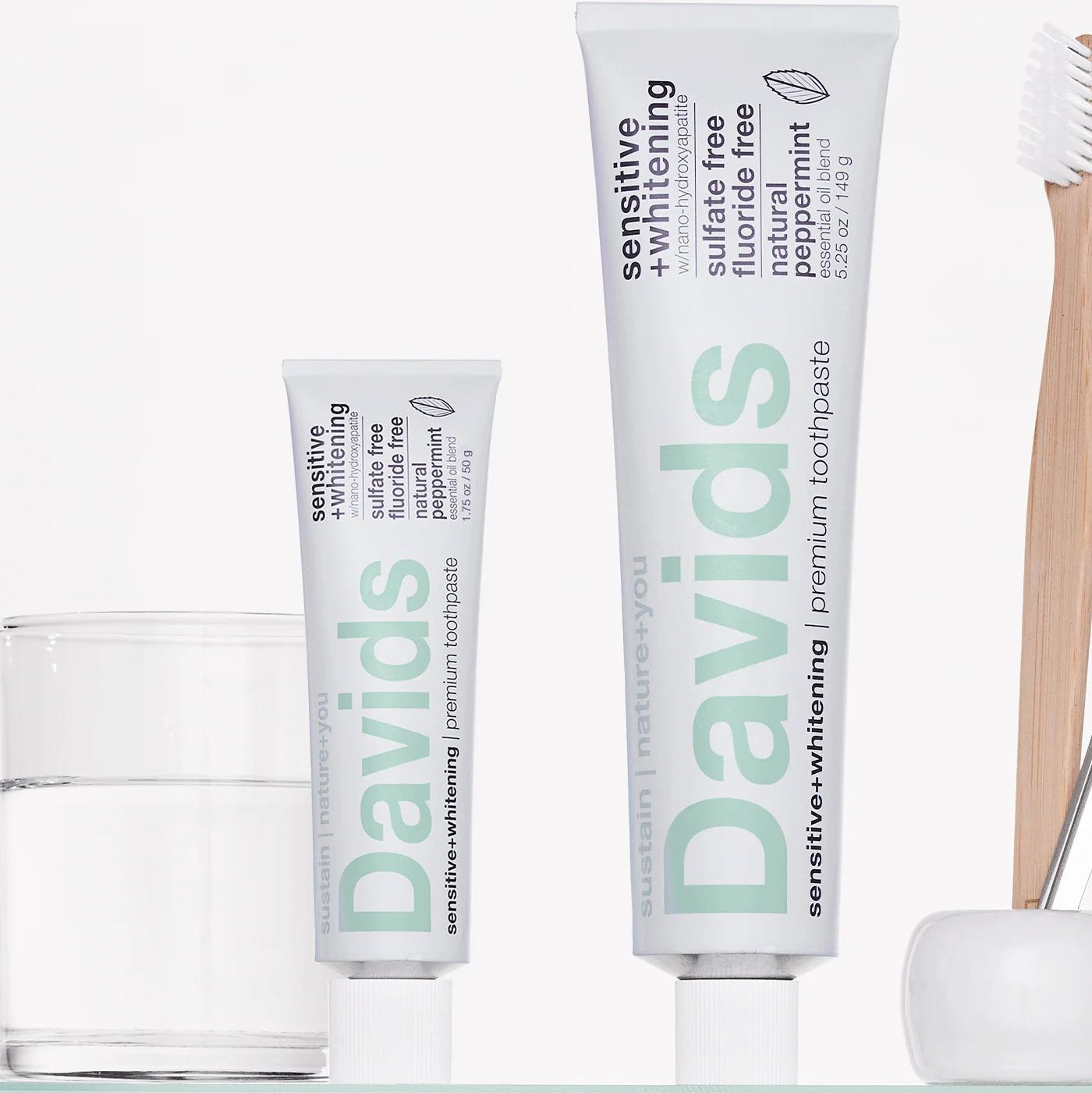 Two tubes of Davids sensitive whitening toothpaste with a glass of water and a wooden toothbrush on a white background.