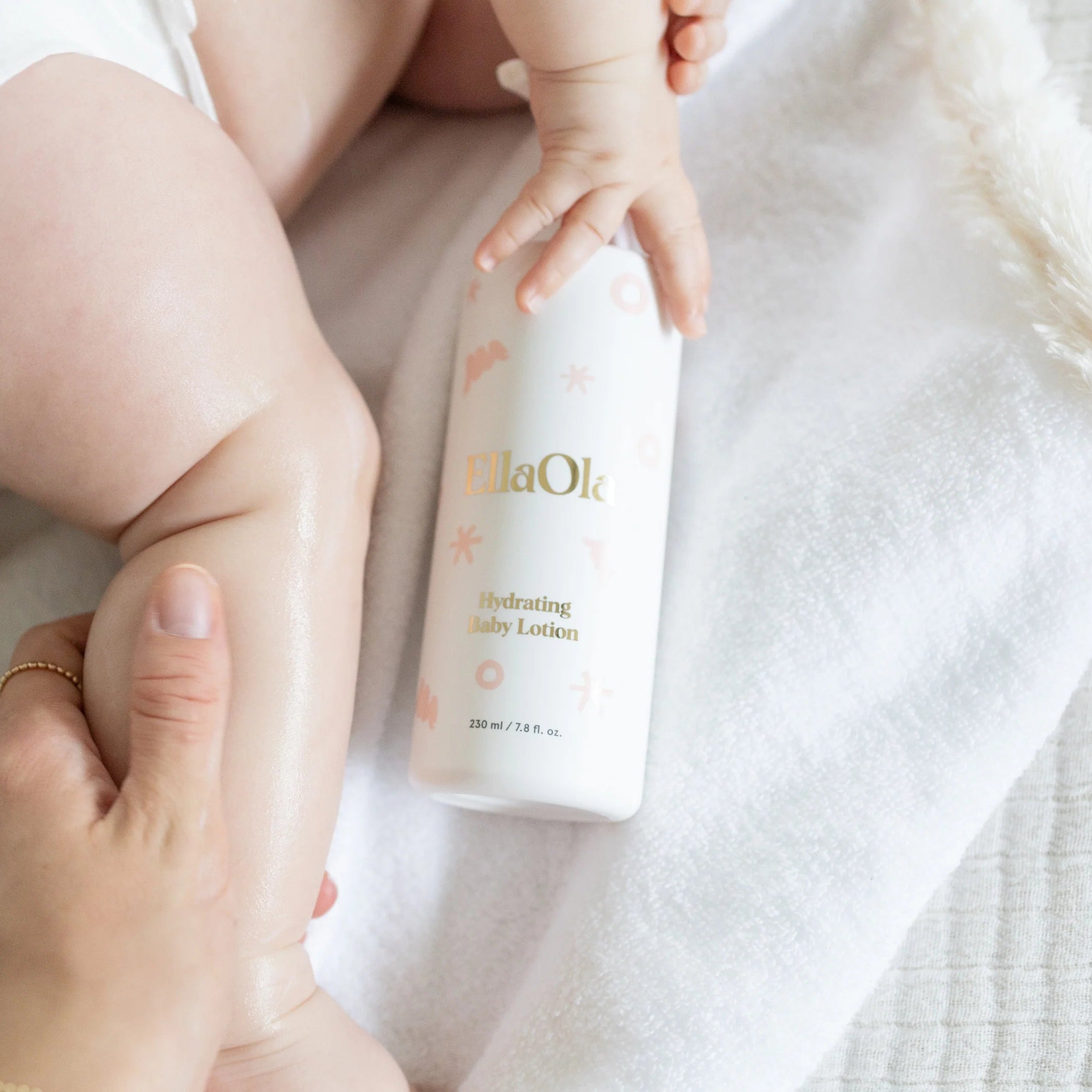 Baby holding a bottle of 'HilaOla' hydrating baby lotion on a soft surface.
