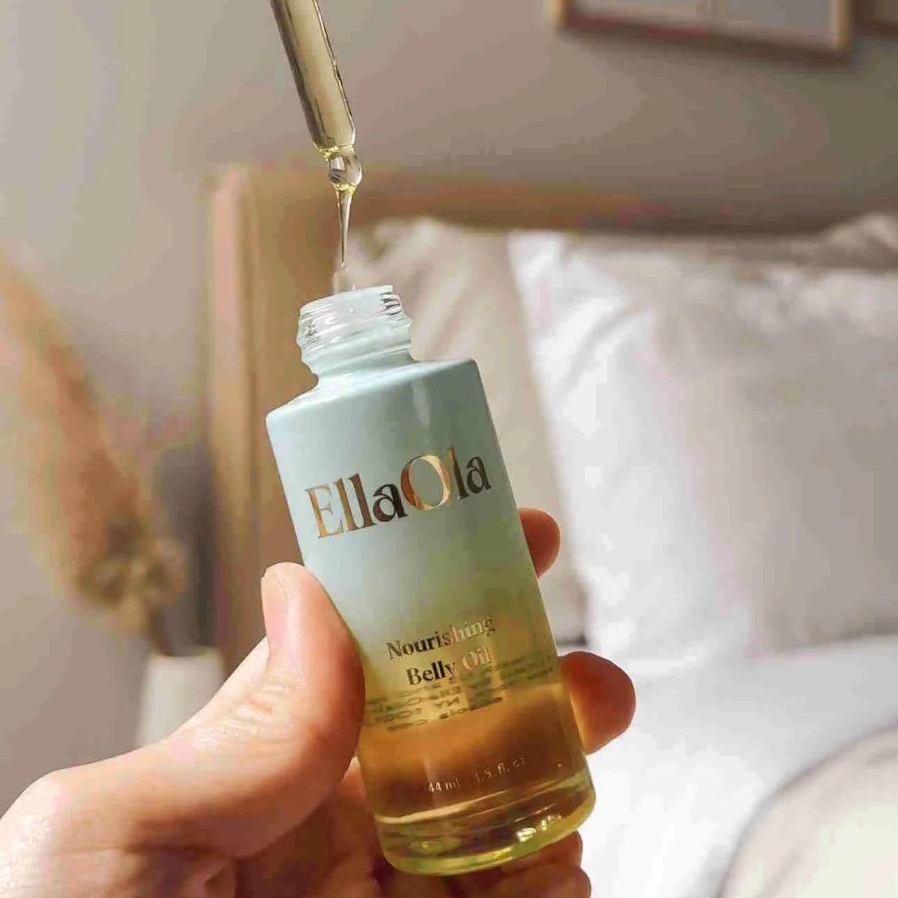EllaOla belly oil bottle with dropper being held, with a blurred indoor background.