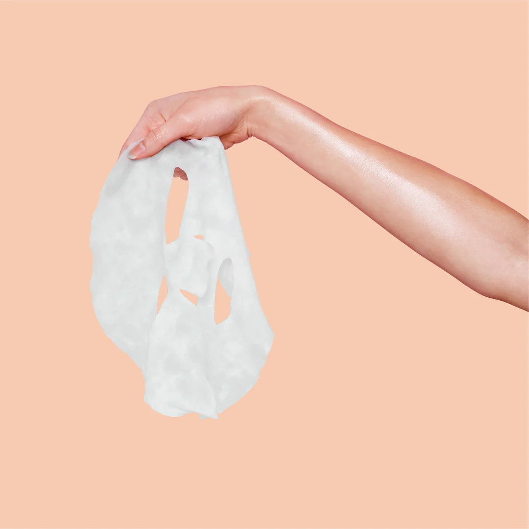 Hand holding a white sheet mask against a peach background