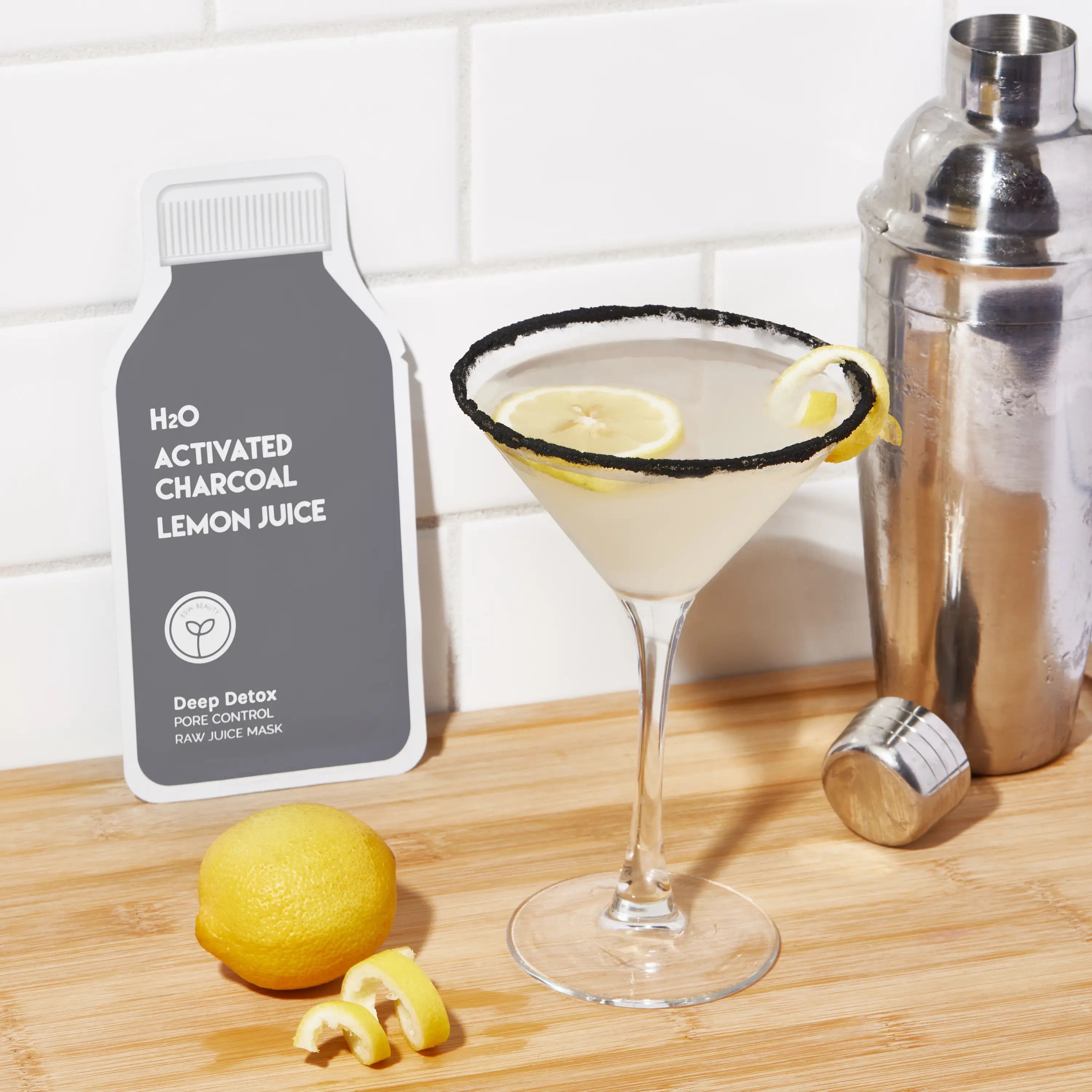 Cocktail with lemon slices, a bottle of H2O Activated Charcoal Lemon Juice, and a cocktail shaker on a wooden surface.
