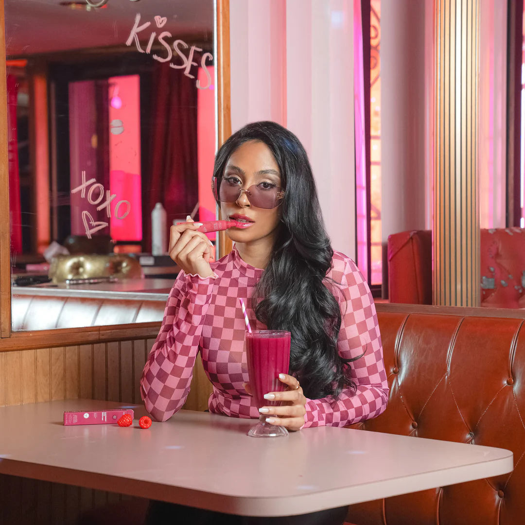 Model applying The Pharmacy Lab ESW Beauty Raspberry Acai Smoothie Enhanced Protection Lip Treatment in a retro café setting with berry smoothie