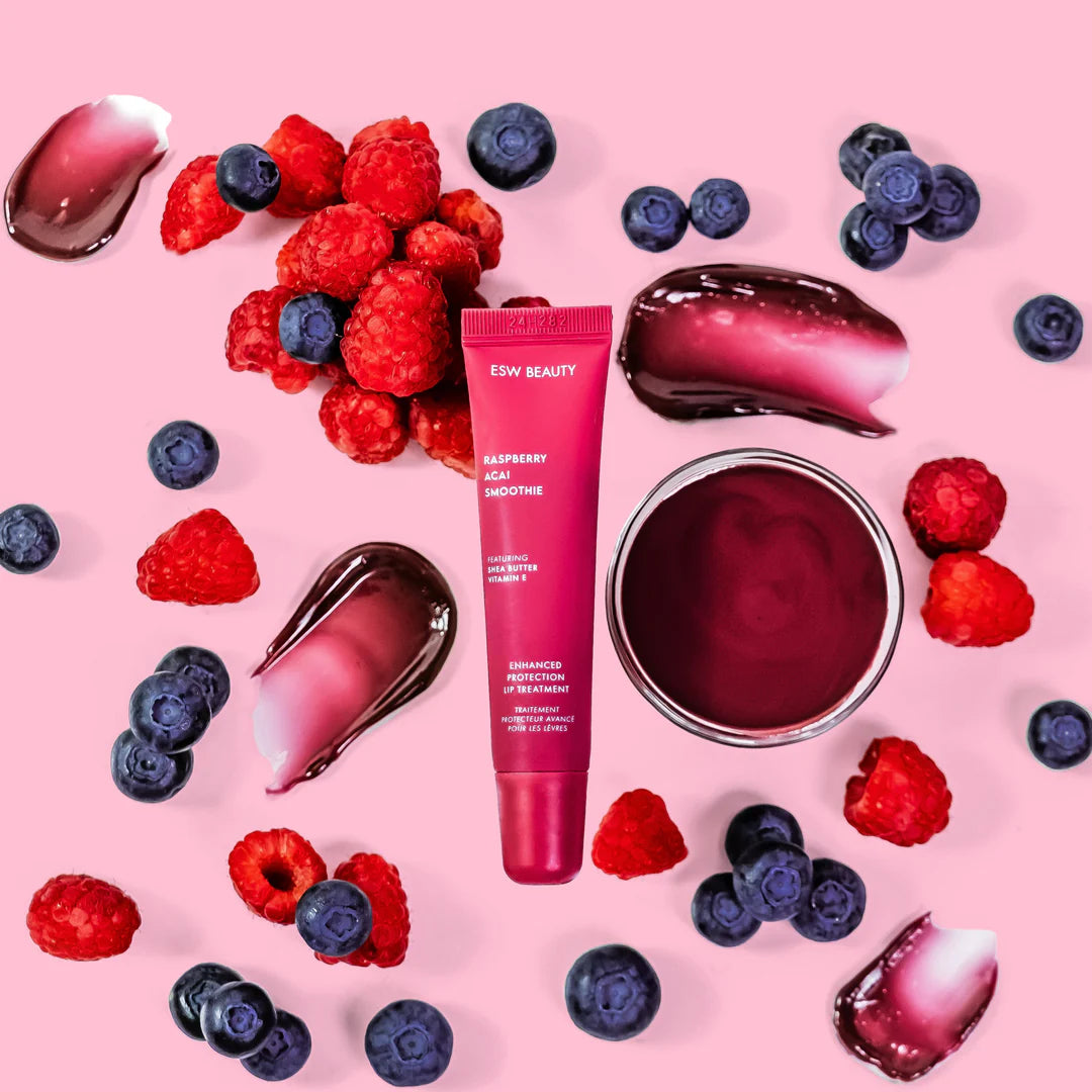 Flat lay of The Pharmacy Lab ESW Beauty Raspberry Acai Smoothie Enhanced Protection Lip Treatment surrounded by raspberries, blueberries, and berry-toned gloss textures