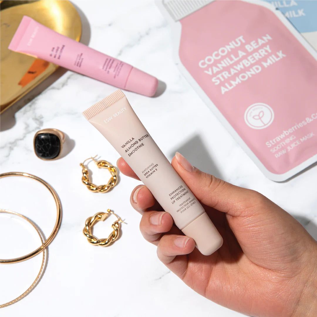 Hand holding a skincare product with beauty items including a pink bottle and gold jewelry on a white surface.