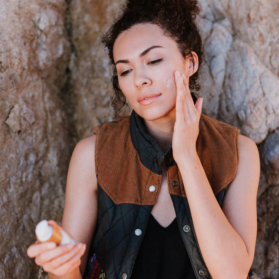 Woman applying sunscreen to her face against a textured stone wall.