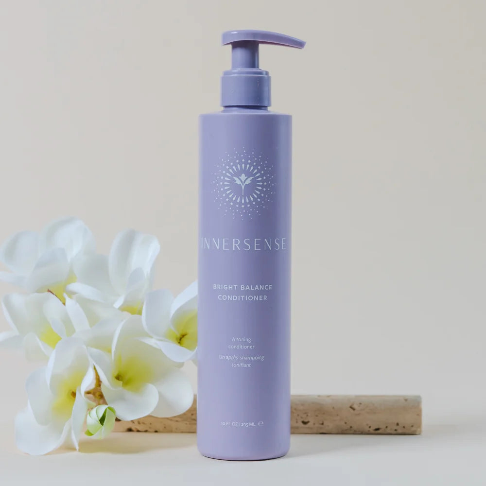 A lavender pump bottle of InnerSense Bright Balance Conditioner stands on a light surface with white plumeria flowers and a cork stick beside it against a warm beige background.