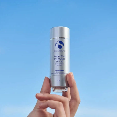 Hand holding a bottle of 'iS Clinical' PerfectTint Powder SPF 40 against a blue background