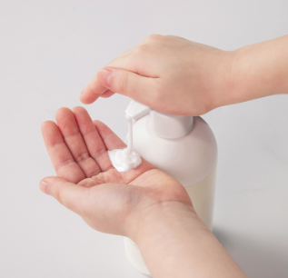 Hand applying a small amount of white cream from a pump bottle onto another hand against a light gray background