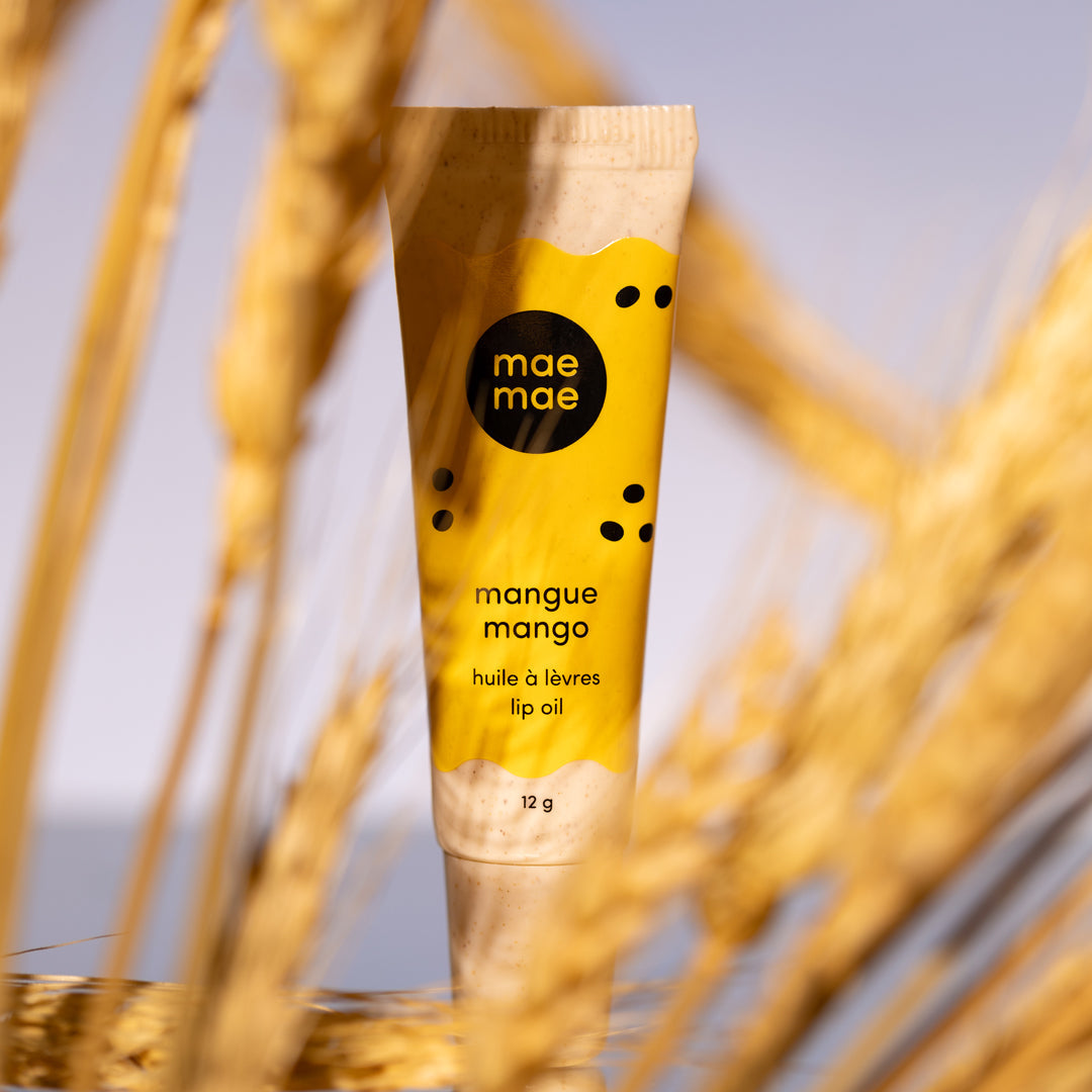 Lip oil tube labeled 'mae mae' with 'mangue mango' flavor against a blurred natural background