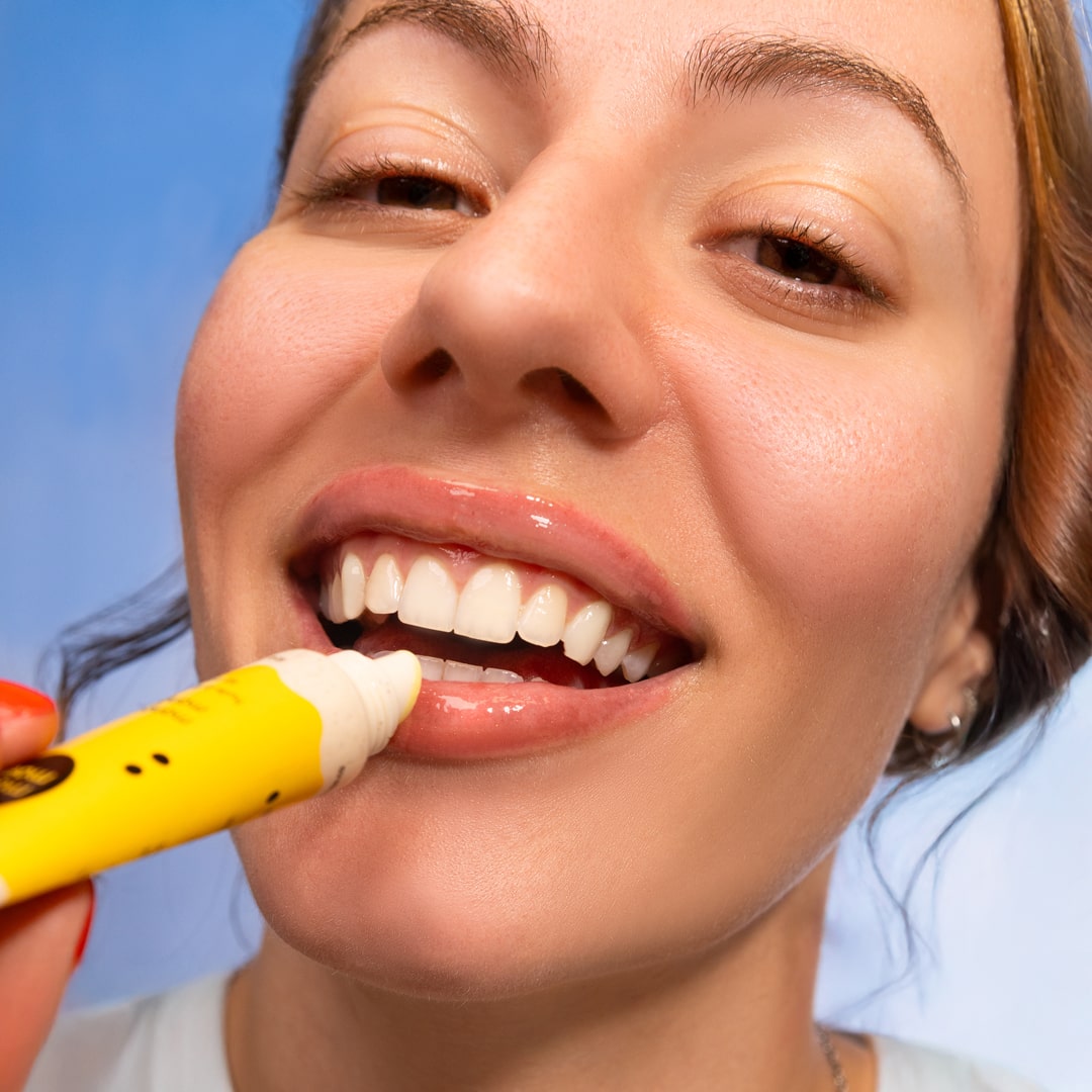 Person applying a white cream to their lips with a yellow applicator against a blue sky background