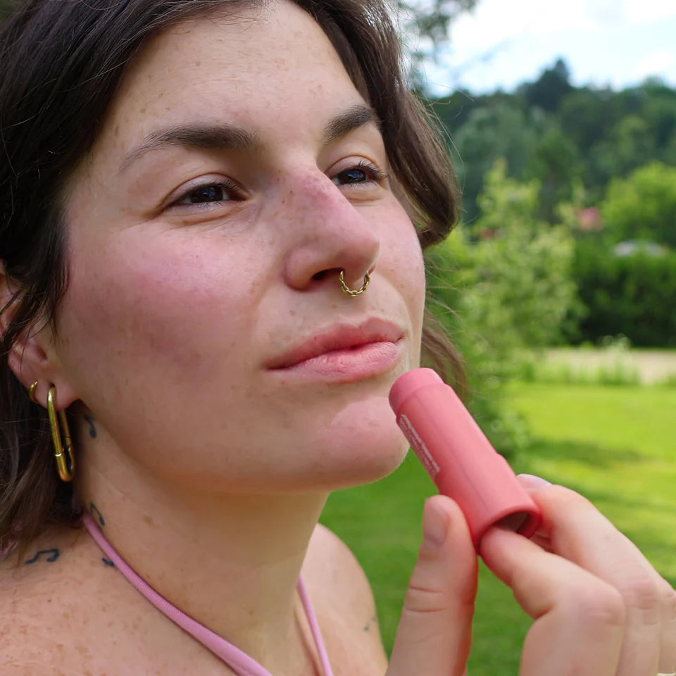 Woman applying pink lipstick outdoors with greenery in the background