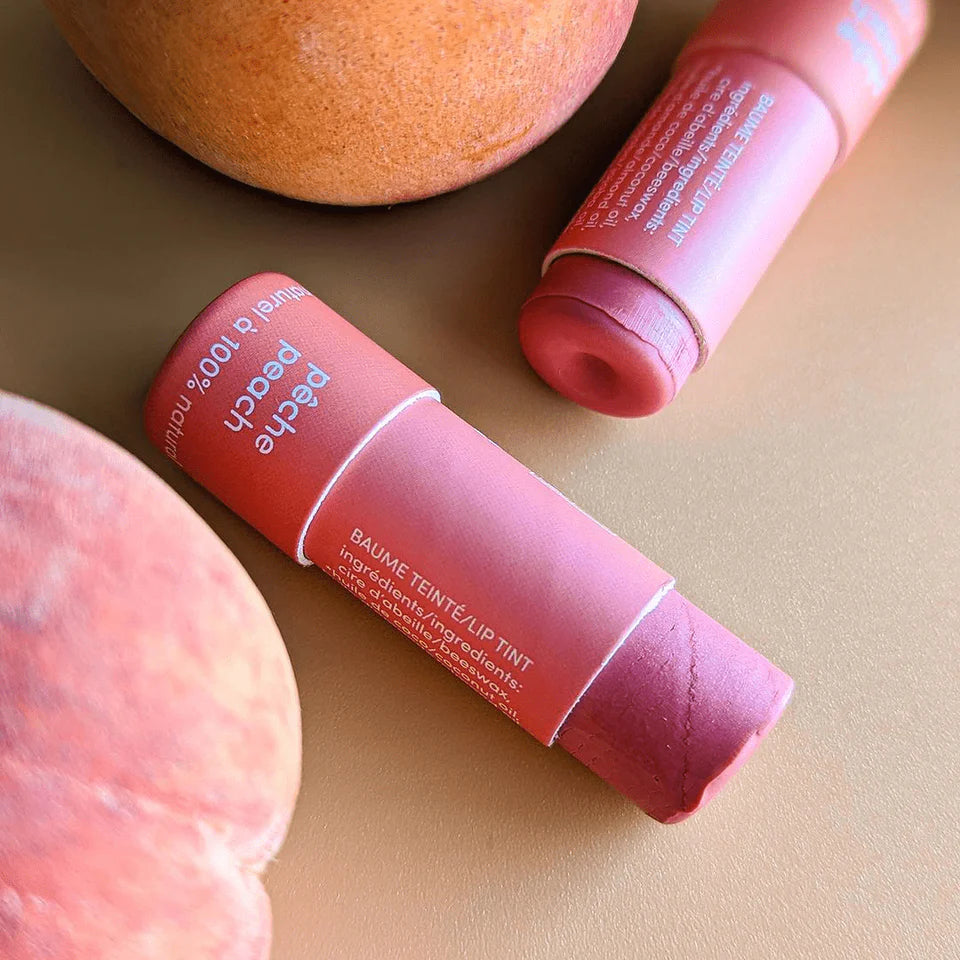 Two pink lip balm tubes on a beige surface with peaches.