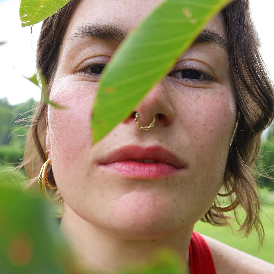Person with a nose ring and hoop earrings peeking through green leaves