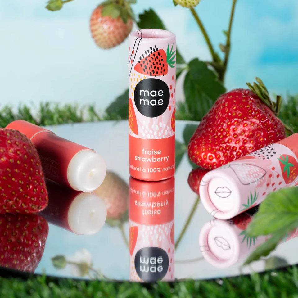Two lip balm tubes with strawberry flavor on a reflective surface with strawberries and greenery in the background.