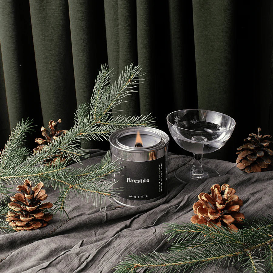 Candle labeled 'fireside' with pine branches and a glass on a dark background
