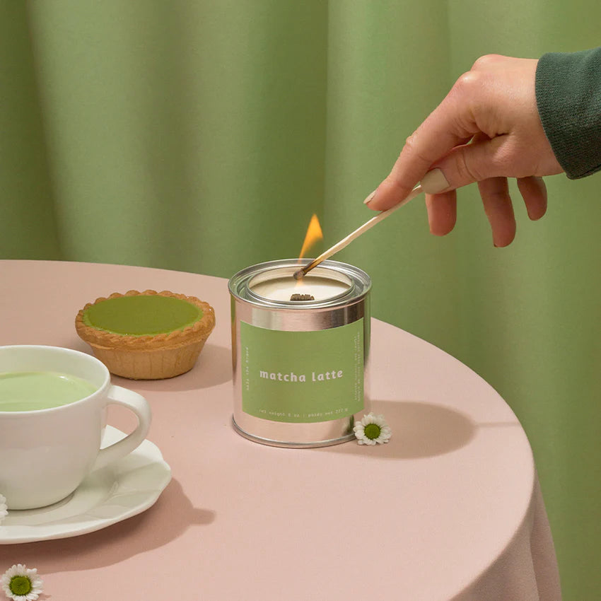 Hand lighting a match next to a 'matcha latte' candle on a table with a green background