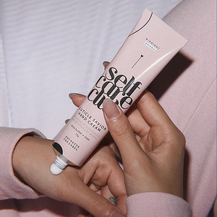 Hand applying hand cream from a pink tube labeled 'self care' with a blurred background.
