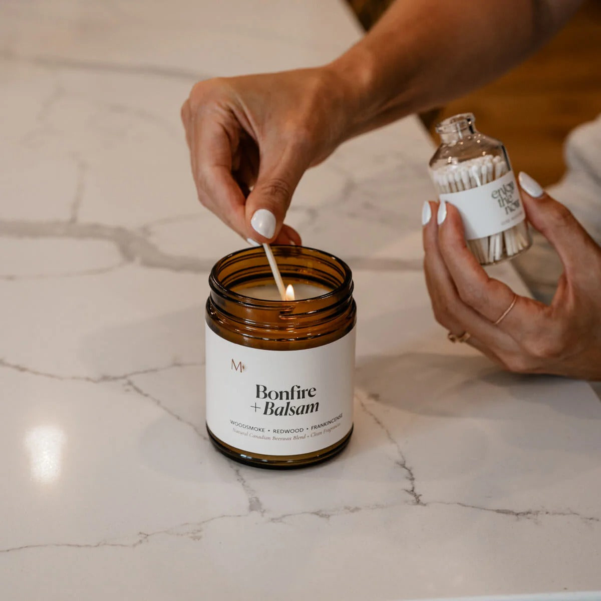 Person lighting a candle labeled 'Bonfire + Balsam' with a match on a marble surface.