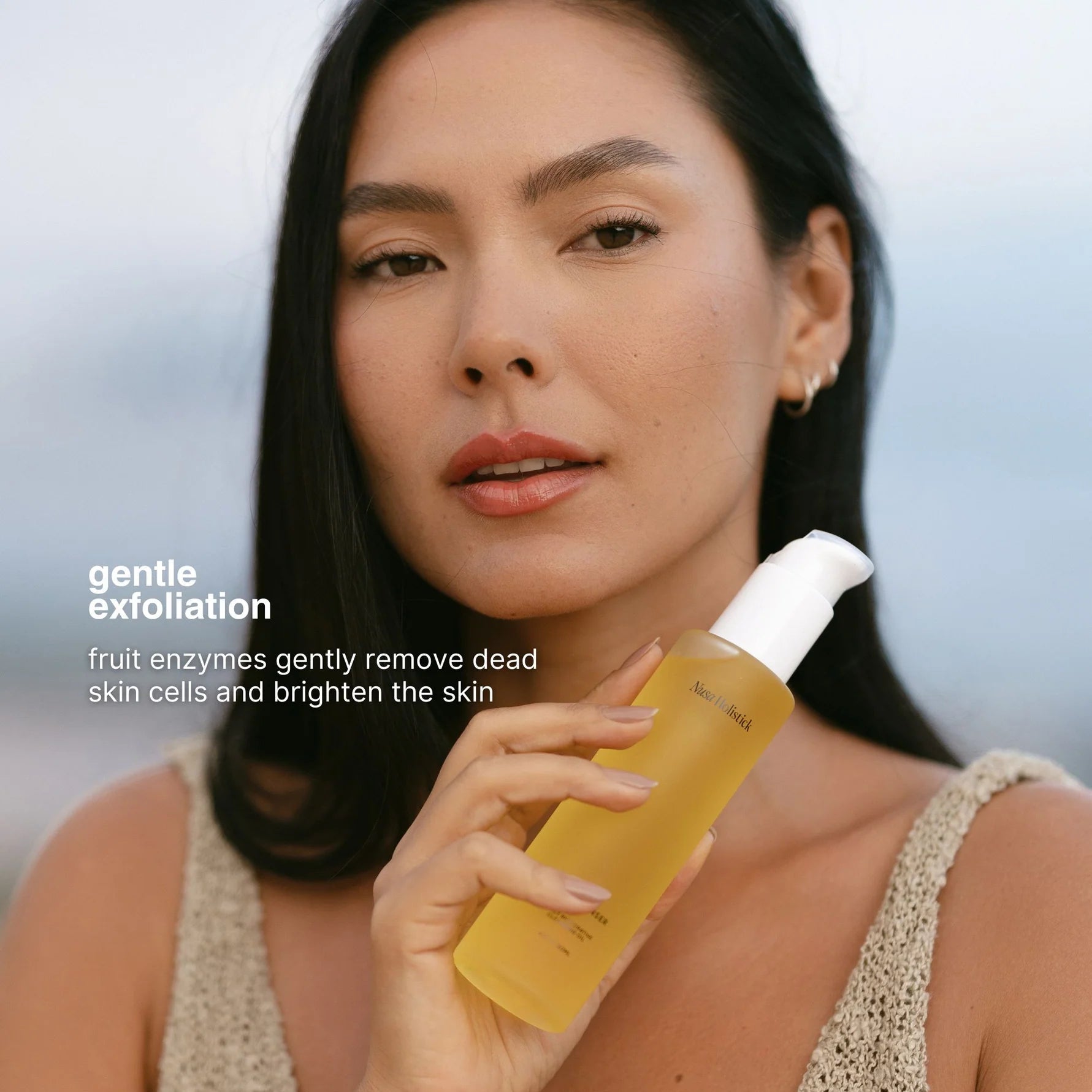 A woman holds a bottle of gentle exfoliating liquid sunscreen against a soft, blurred outdoor background. The bottle is filled with a golden-yellow liquid and has a white pump dispenser.