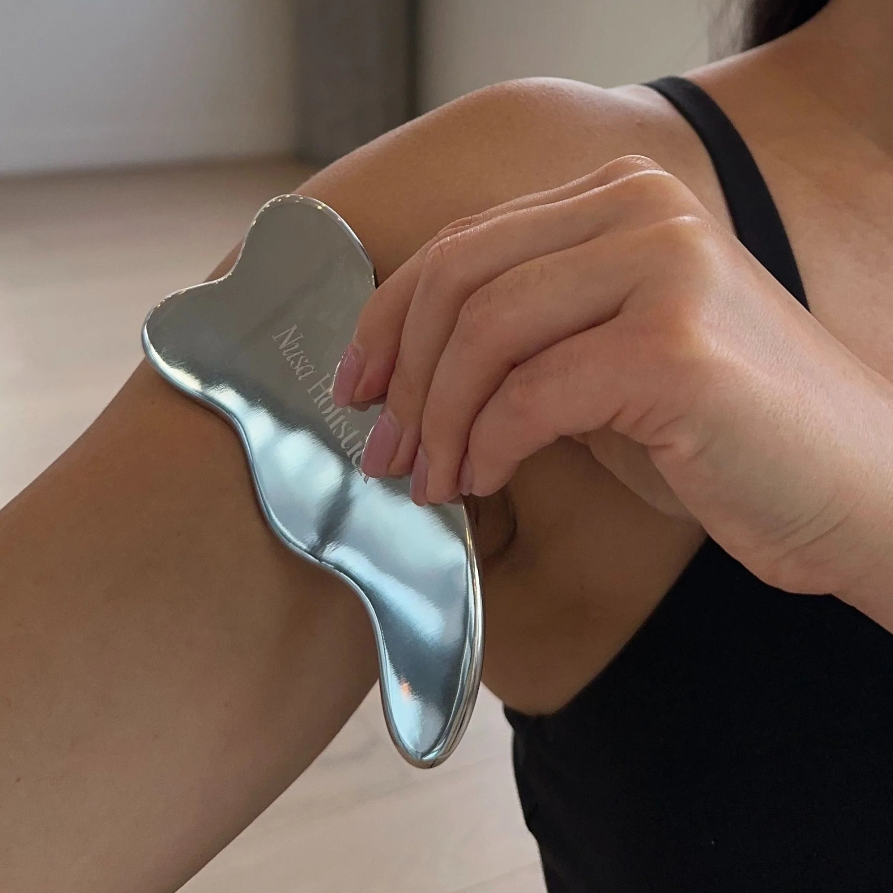 Person using a silver gua sha tool on their arm in a bright room.