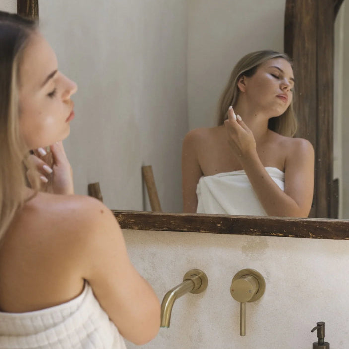 Woman in a towel looking at herself in a mirror in a bathroom.