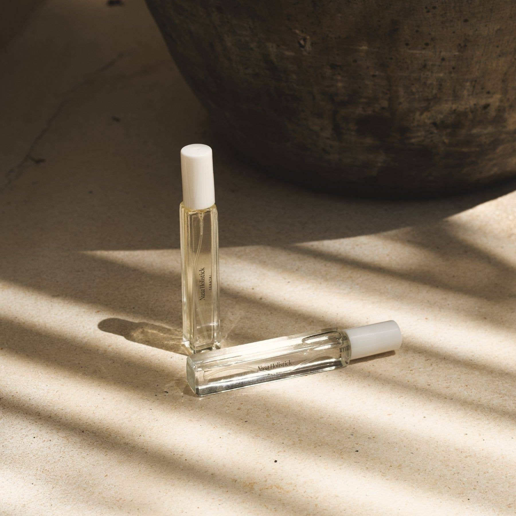 Two clear glass rollerball bottles with white caps on a textured surface with shadows.