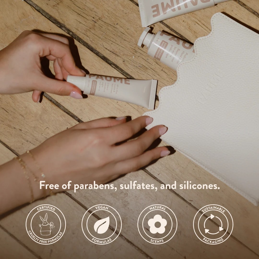 Hand holding a tube of cream on a wooden floor with product packaging in the background, featuring text about product ingredients.