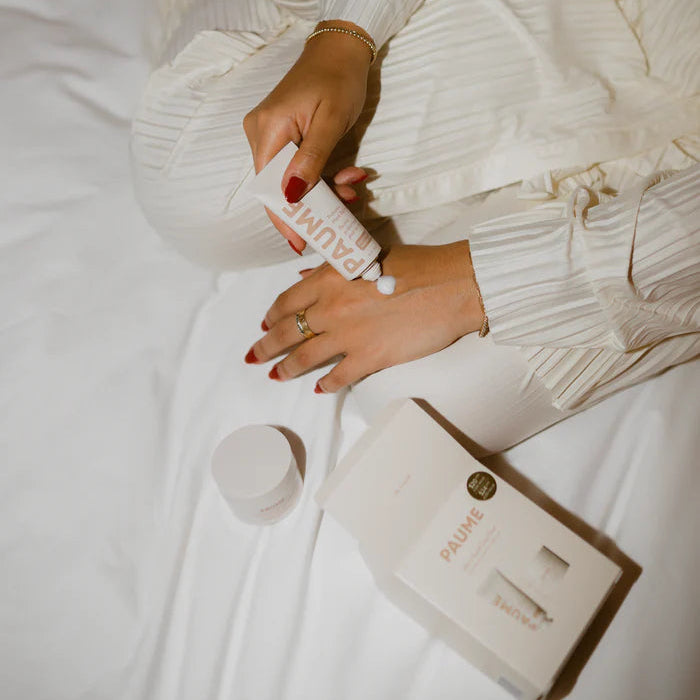 Person holding a bottle of 'Nuxe' product on a bed