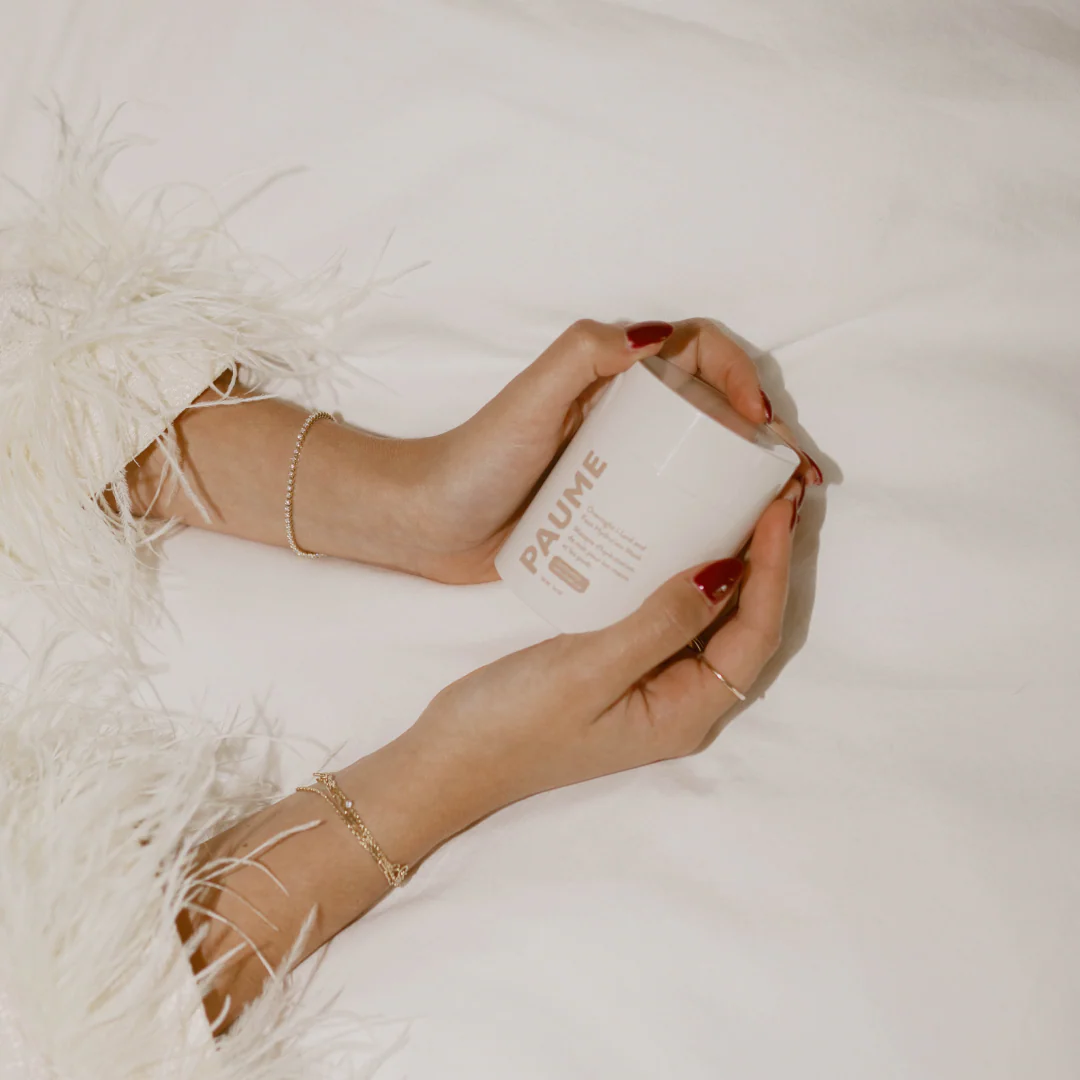 Person holding a white container labeled 'PAUME' with feet wearing strappy sandals on a soft surface.