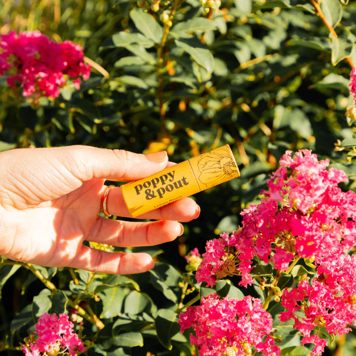 Hand holding a yellow product labeled 'poppy & pout' among pink flowers