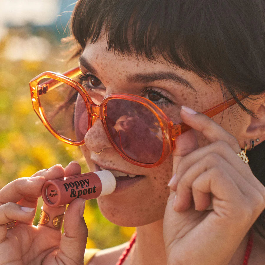 Woman applying lip balm with 'poppy & pout' packaging outdoors.
