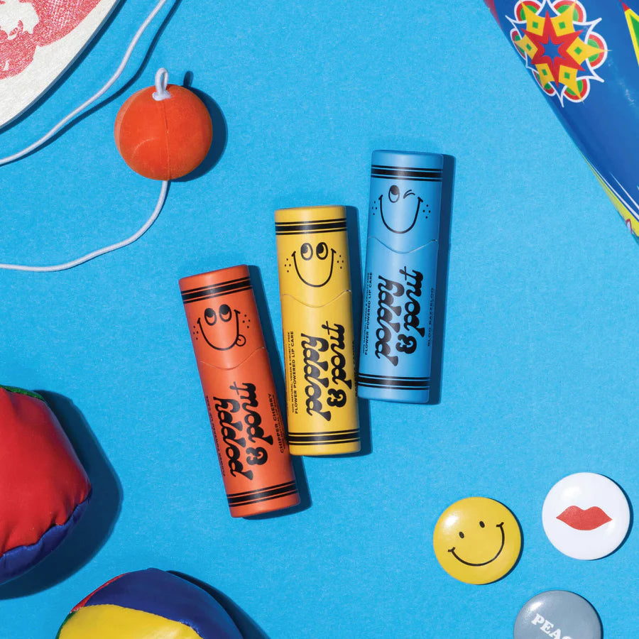 Three colorful lip balm tubes with smiley faces on a blue background, surrounded by small toys and buttons.