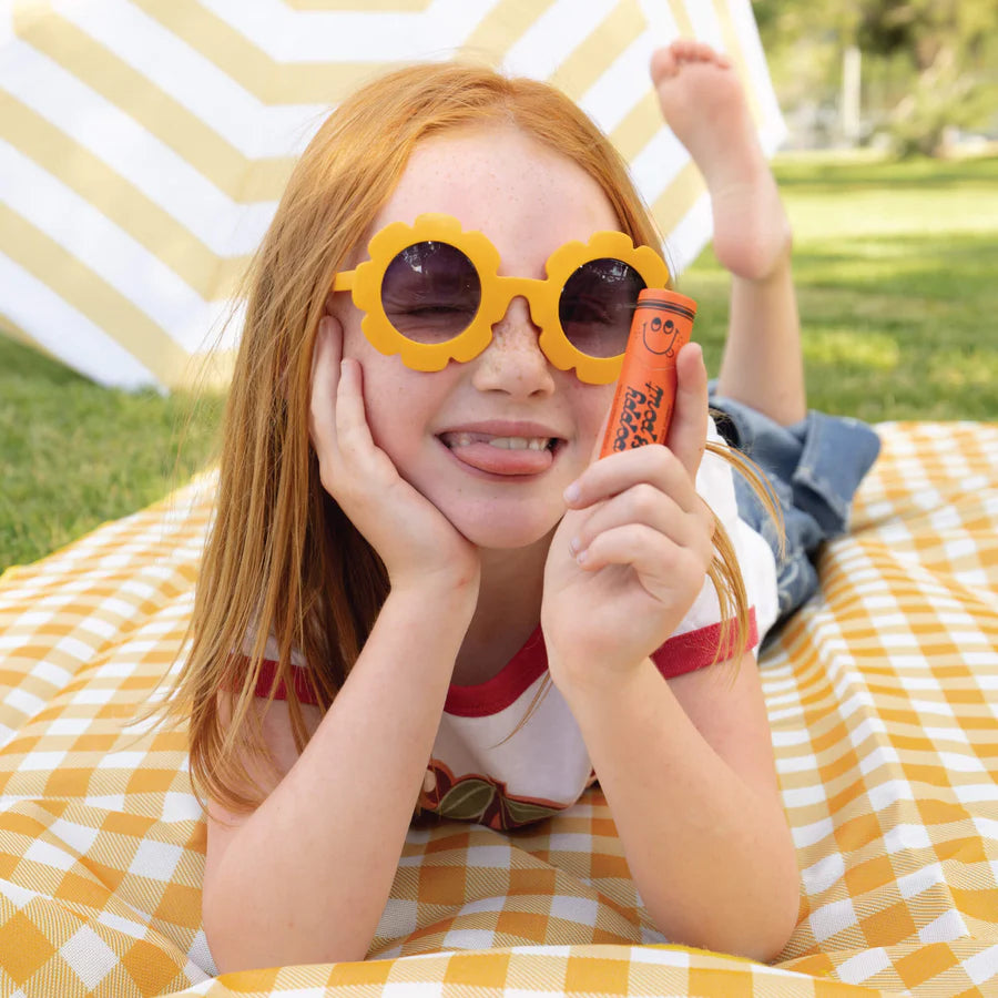 Child wearing yellow sunglasses and holding a red object on a checkered blanket outdoors.