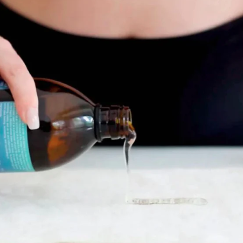 Person pouring a liquid from a brown bottle with a blue label onto a white surface.